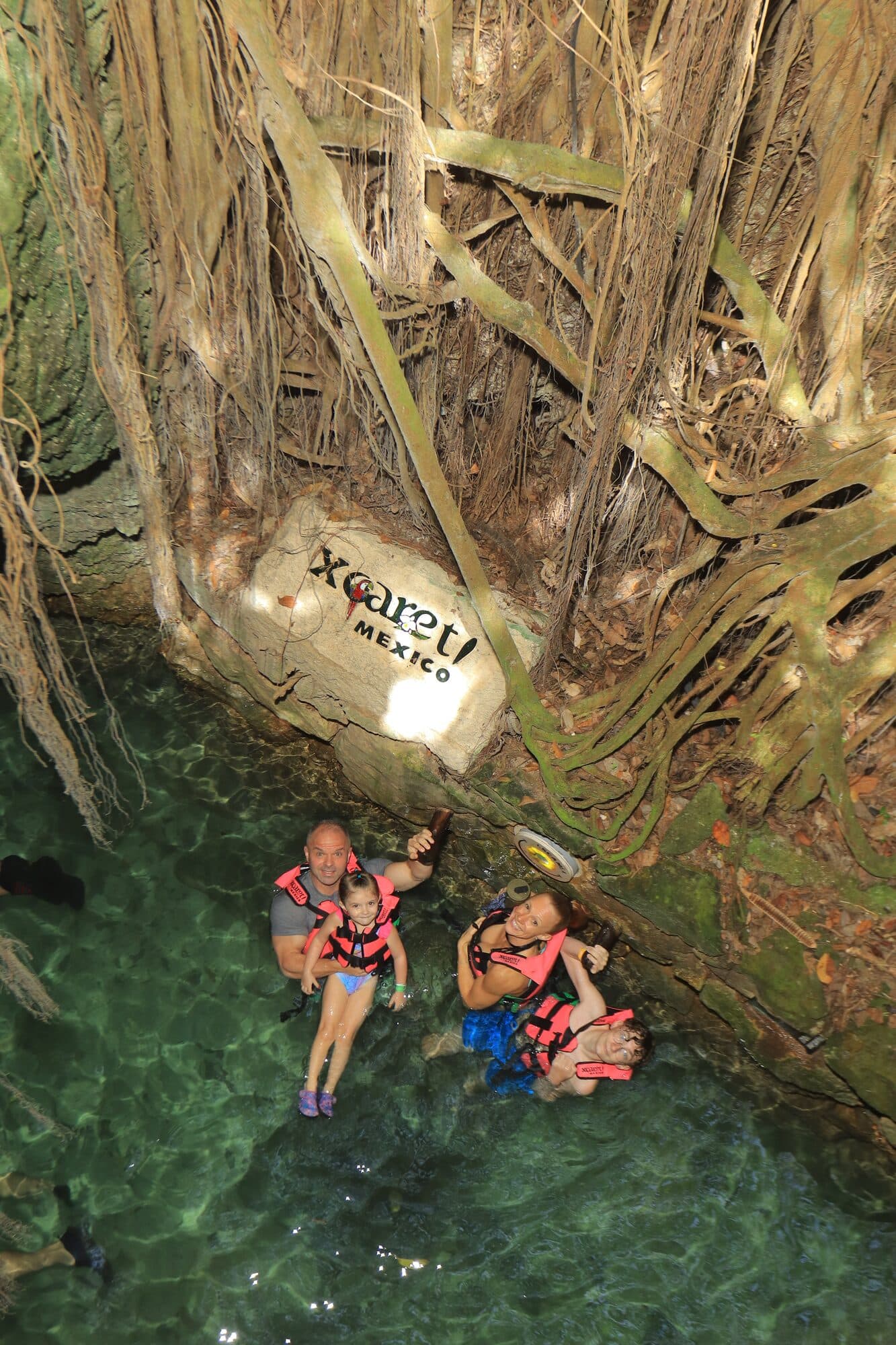 A family floating the Xcaret underground river in life jackets beneath hanging jungle roots