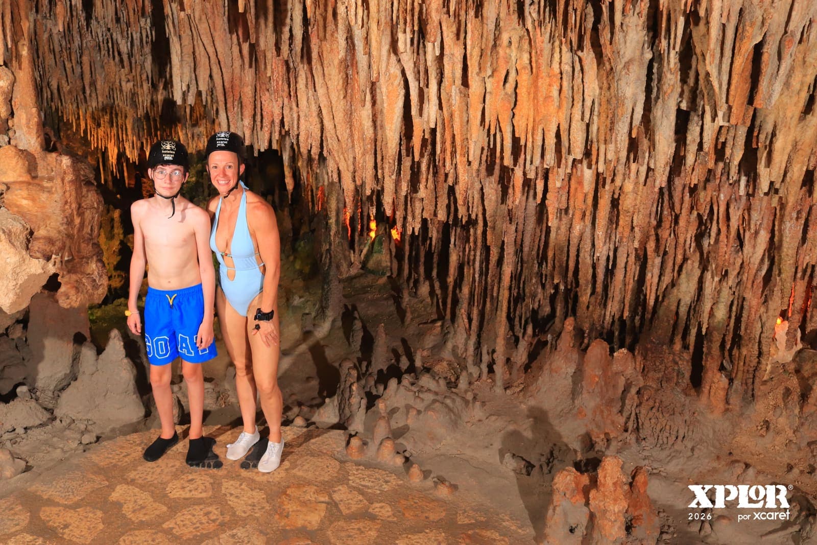 Two visitors in safety helmets standing beneath dense stalactites in an Xplor cave